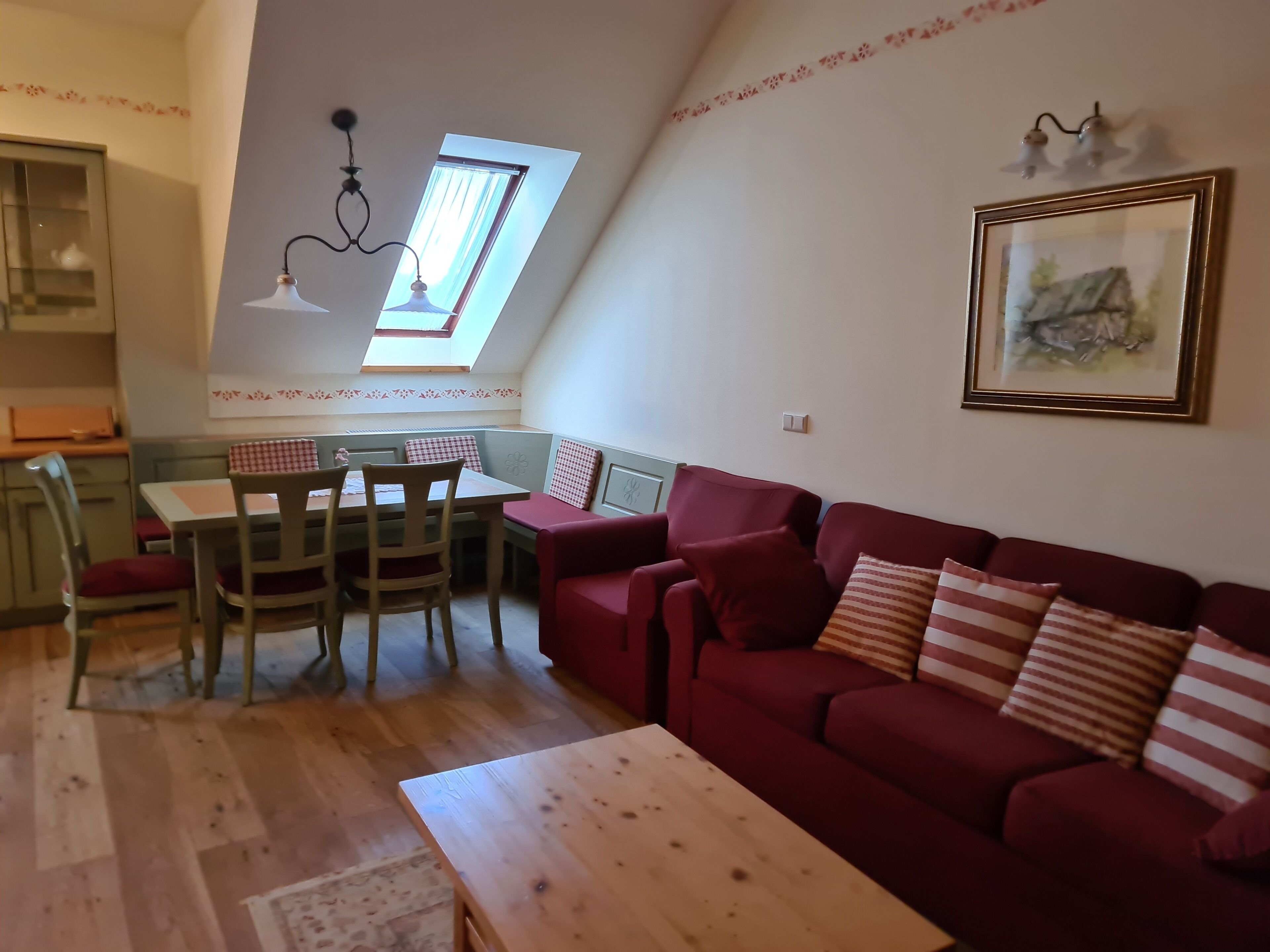A cozy living room with a red sofa, wooden coffee table, and a dining area with a table and chairs. A skylight provides natural light, and a framed picture adorns the wall.
