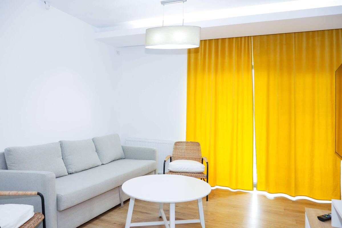 Ein modernes Wohnzimmer mit einem grauen Sofa, einem weißen runden Tisch, einem Korbstuhl und einem gelben Vorhang.