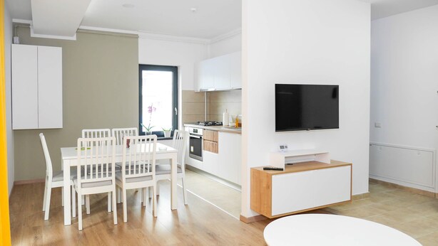 Een moderne keuken met witte kasten, een eethoek met witte stoelen en een flatscreen televisie aan de muur.