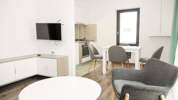 Una habitación moderna y luminosa, con televisión de pantalla plana, una mesa de comedor con sillas, una zona de cocina y un ventanal grande.