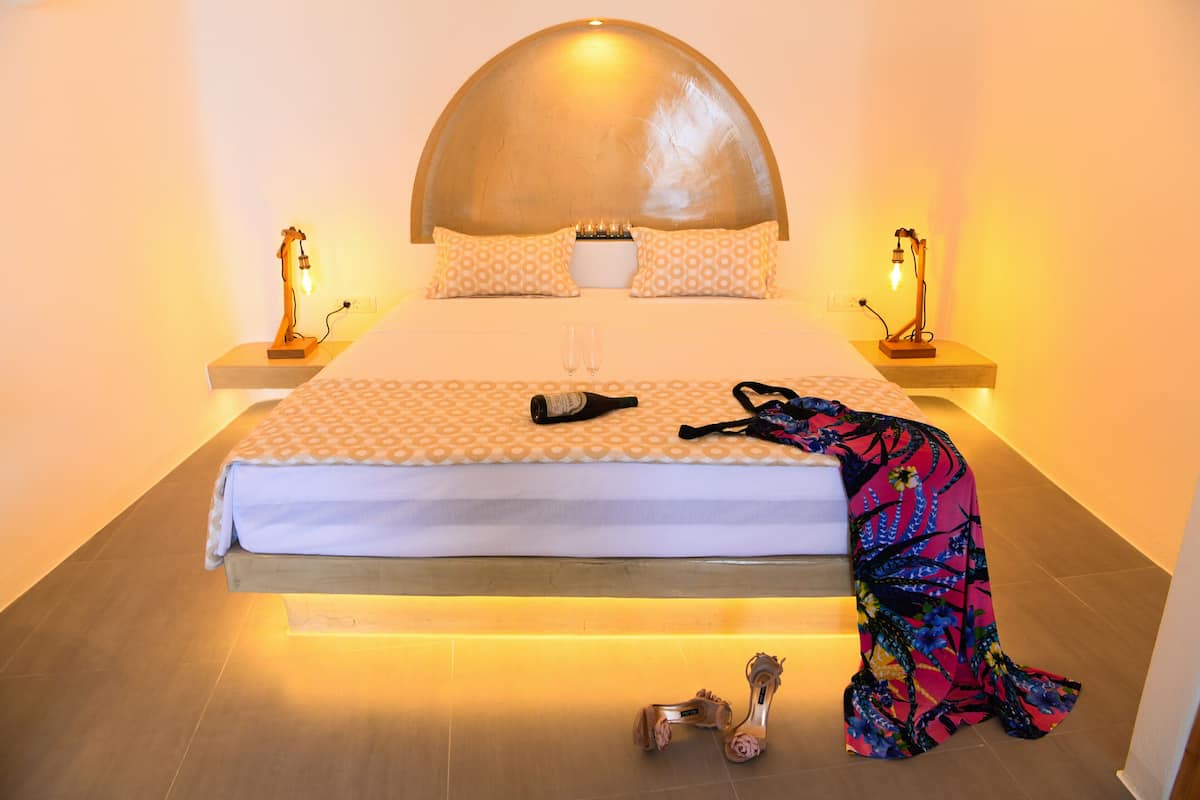 A bed with a white and patterned bedspread, two bedside lamps, a pair of shoes, and a colorful blanket on the floor.