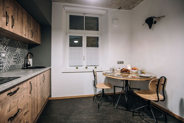 A kitchen with wooden cabinets, a window with white curtains, a round table with chairs, and a wall-mounted light fixture. - BeCycle Sustainable Stay and Travel (Maribor)