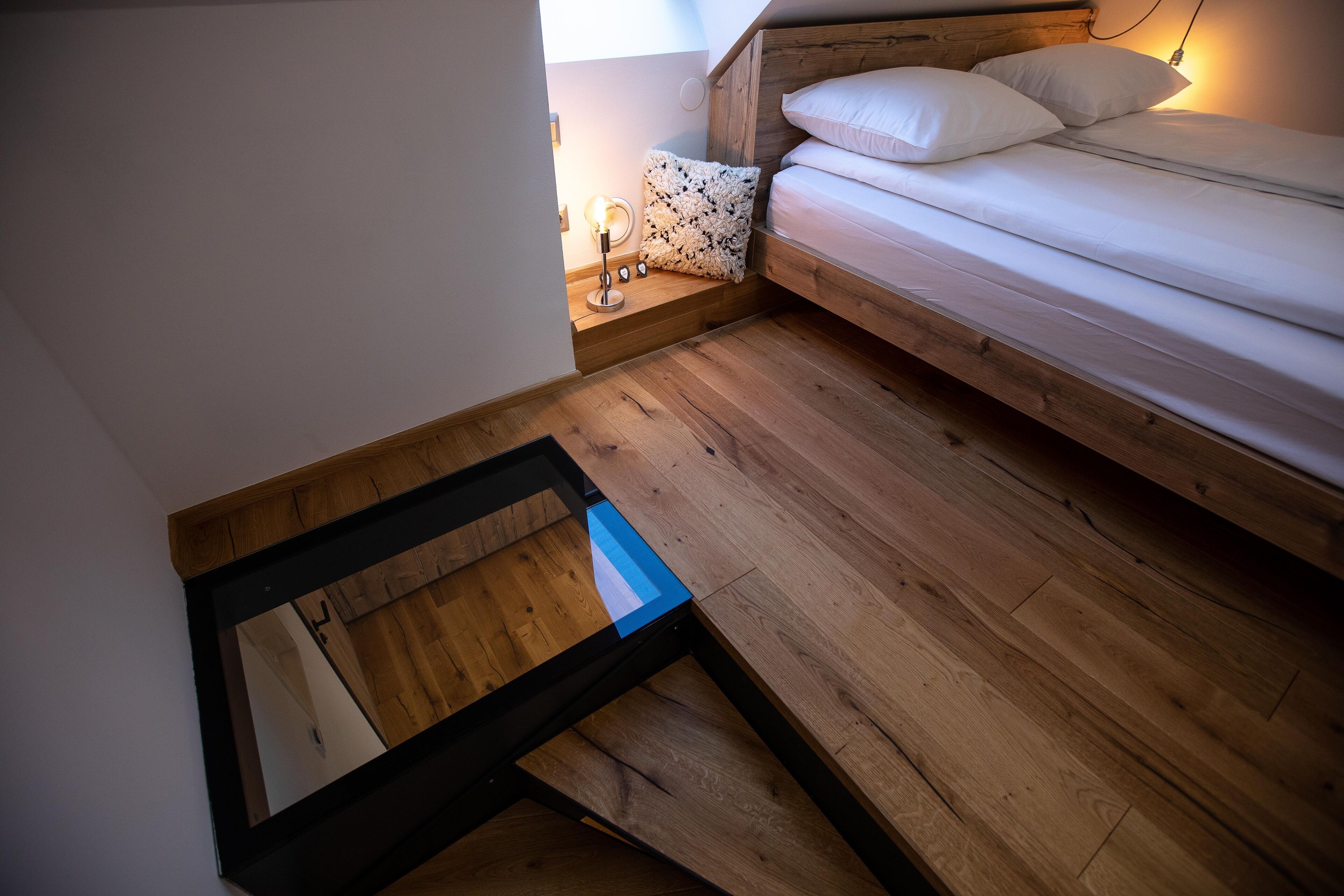 Un dormitorio con suelo de madera, una cama con ropa blanca y una mesita de noche con lámpara.