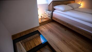 Un dormitorio con suelo de madera, una cama con ropa blanca y una mesita de noche con lámpara.