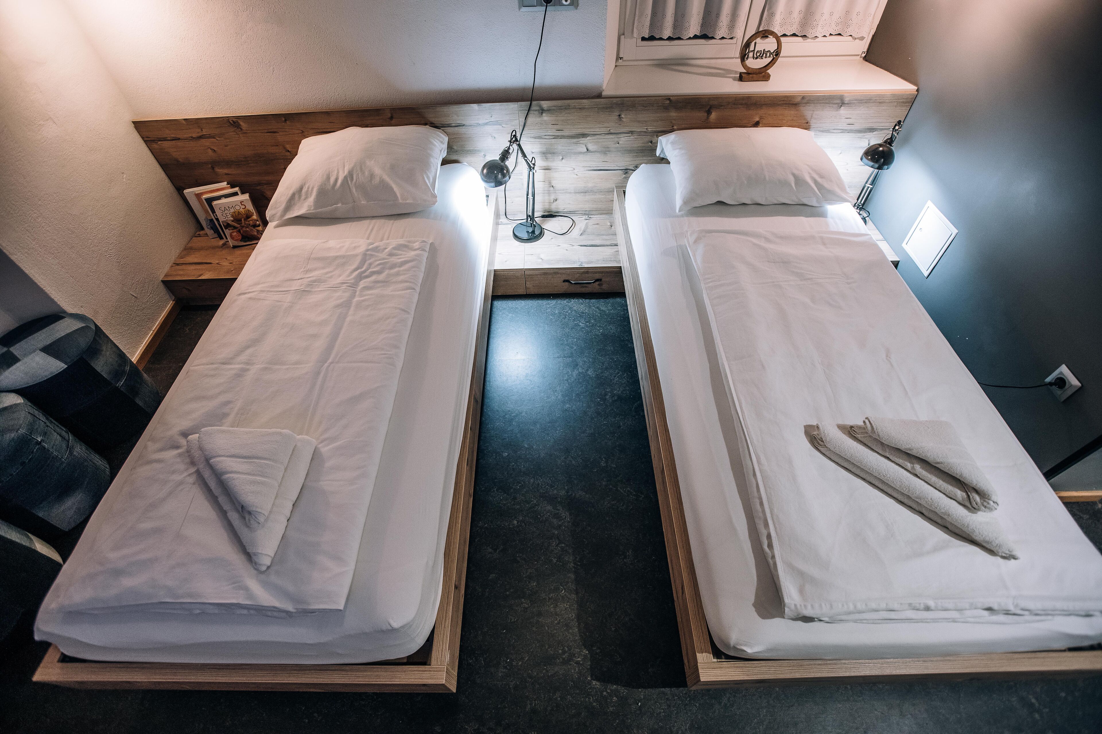 Habitación doble con dos camas individuales, cabecera de madera, una mesita de noche con lámpara y una ventana con cortinas.