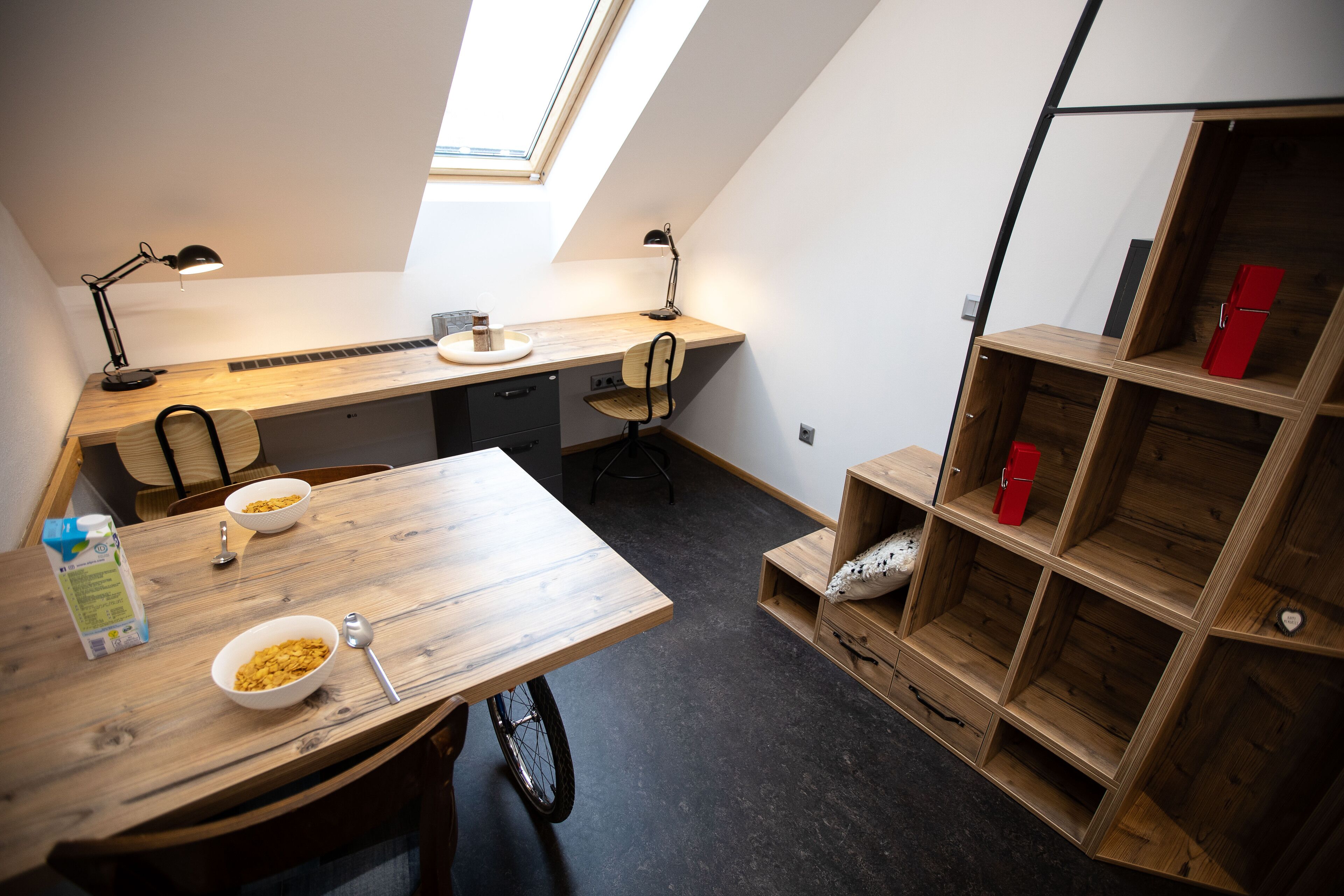 Una habitación compacta y moderna con techo inclinado, un escritorio de madera con dos sillas, una pequeña kitchenette y estanterías empotradas con un electrodoméstico rojo.