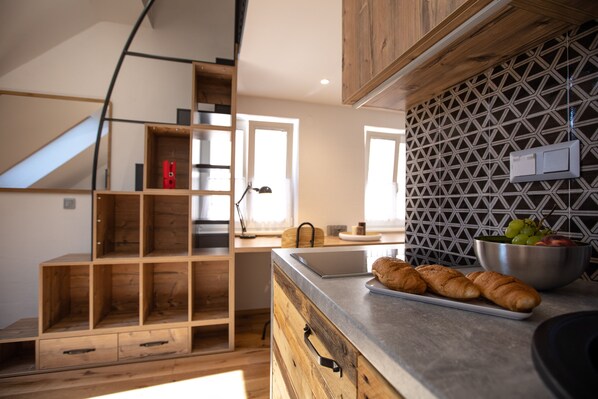 A modern kitchen with wooden cabinetry, a built-in shelf unit, and a countertop with bread and a bowl of fruit.