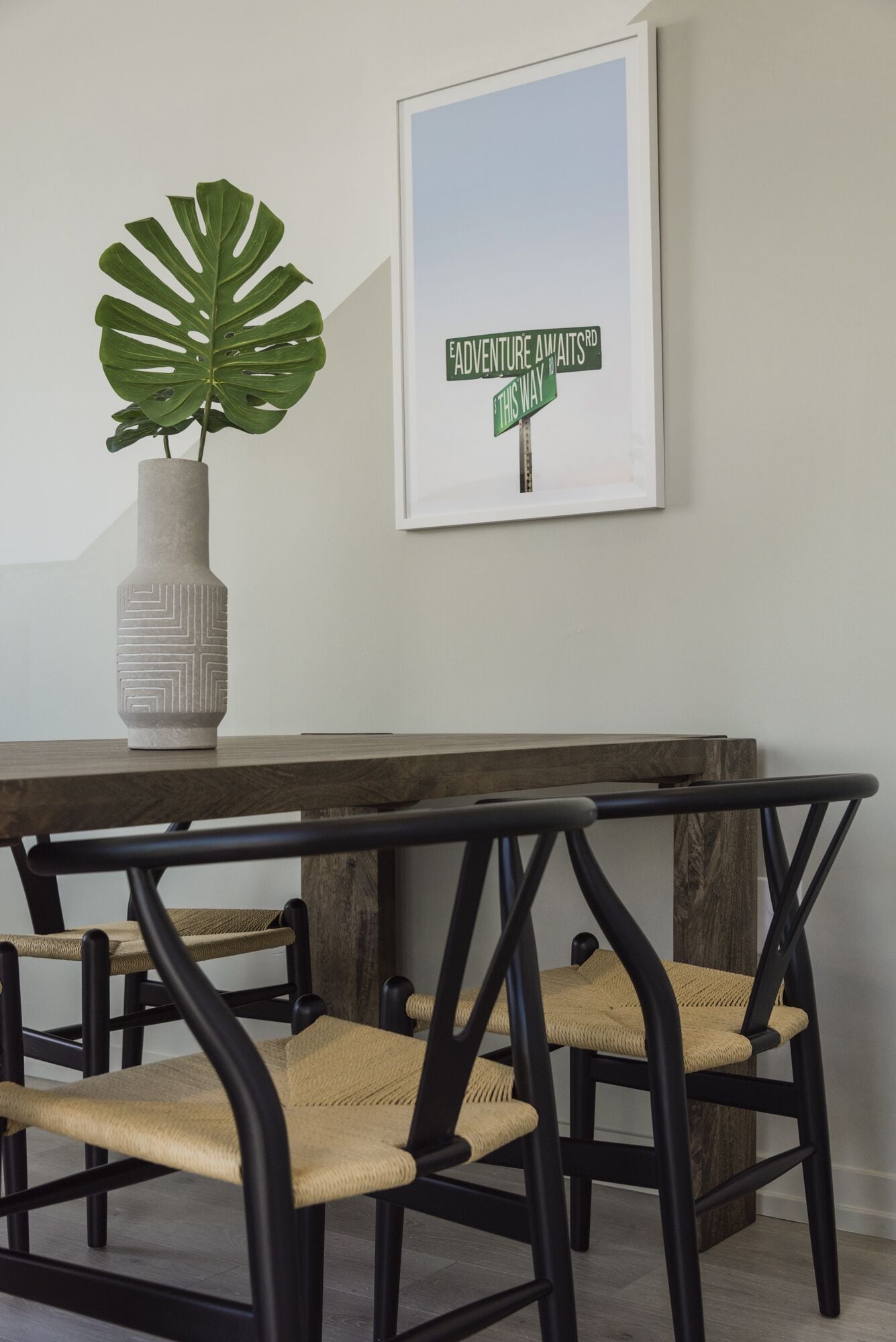 Een eetruimte met een houten tafel, vier zwarte stoelen met gevlochten zittingen, een grote groene plant in een decoratieve vaas en een geïnspireerd citaat in een lijst op de muur.
