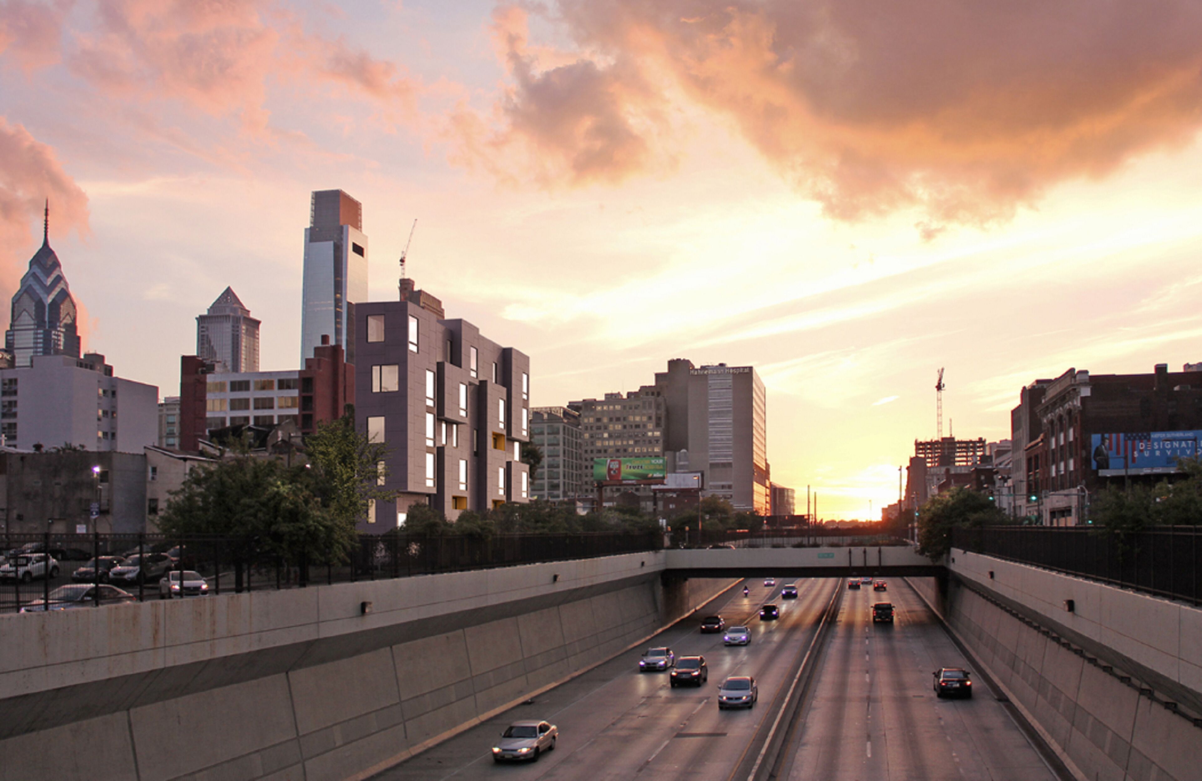 A multi-lane highway with vehicles, surrounded by modern buildings and a sunset sky.
