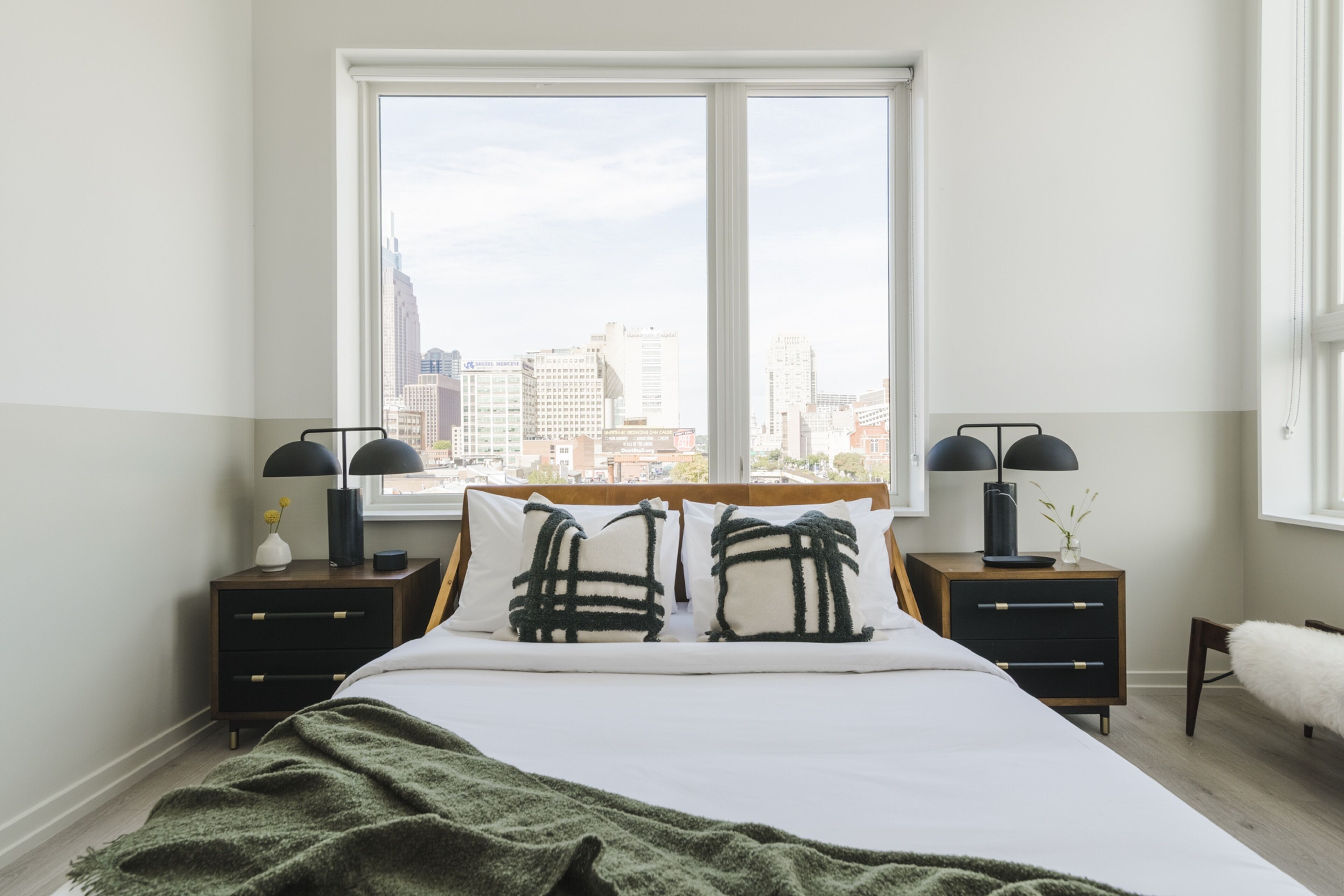 A bedroom with a large window, a bed with two black and white pillows, and two bedside tables with lamps.