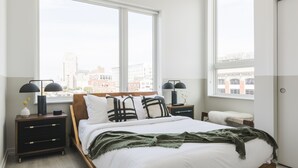 A modern bedroom with a large bed, two bedside tables, and a view of a cityscape through large windows. - Abode Philadelphia - Center City Convention Center (Philadelphia)