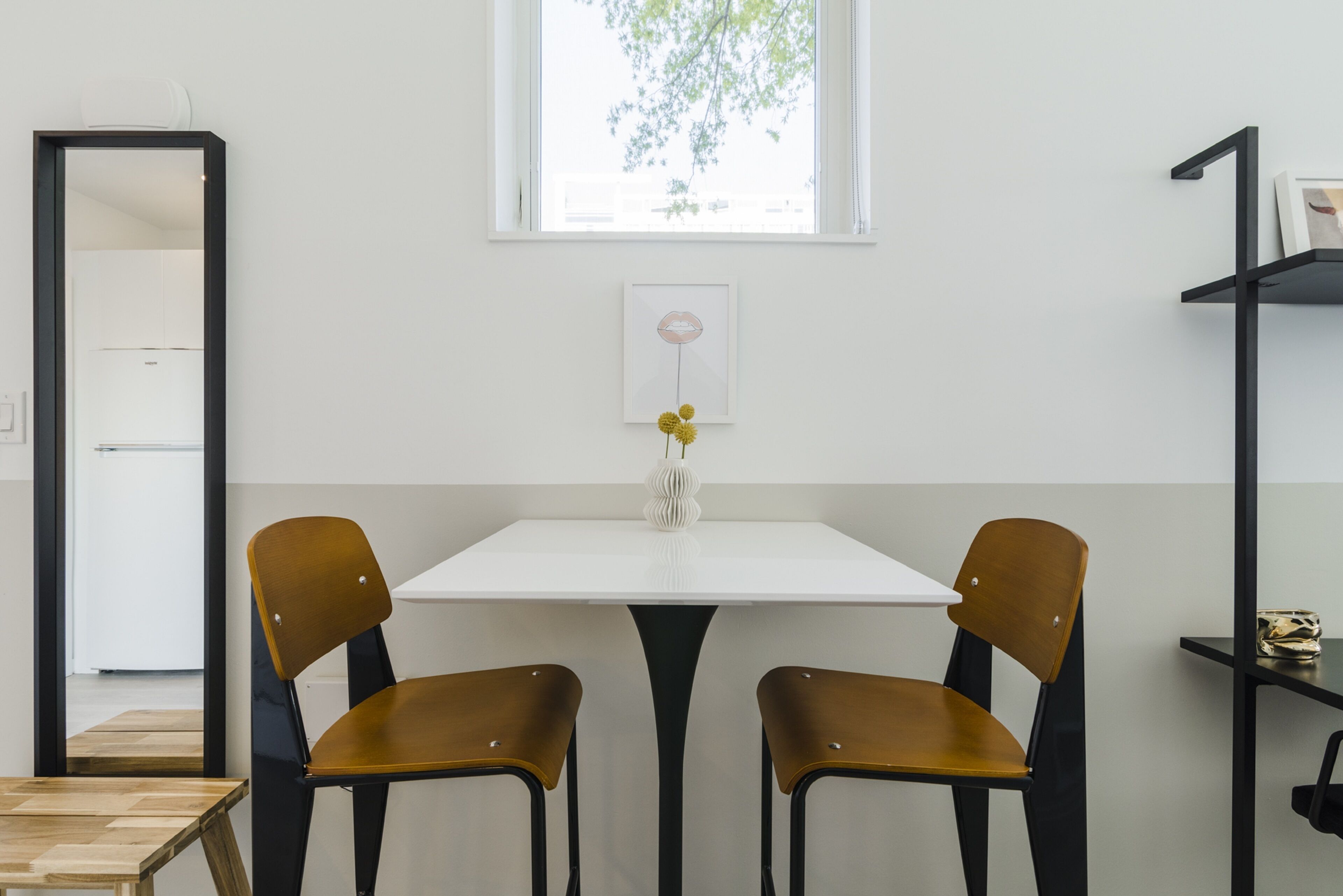 A minimalist room with a white table, two brown chairs, a vase with yellow flowers, a framed picture on the wall, and a window with a view of trees.