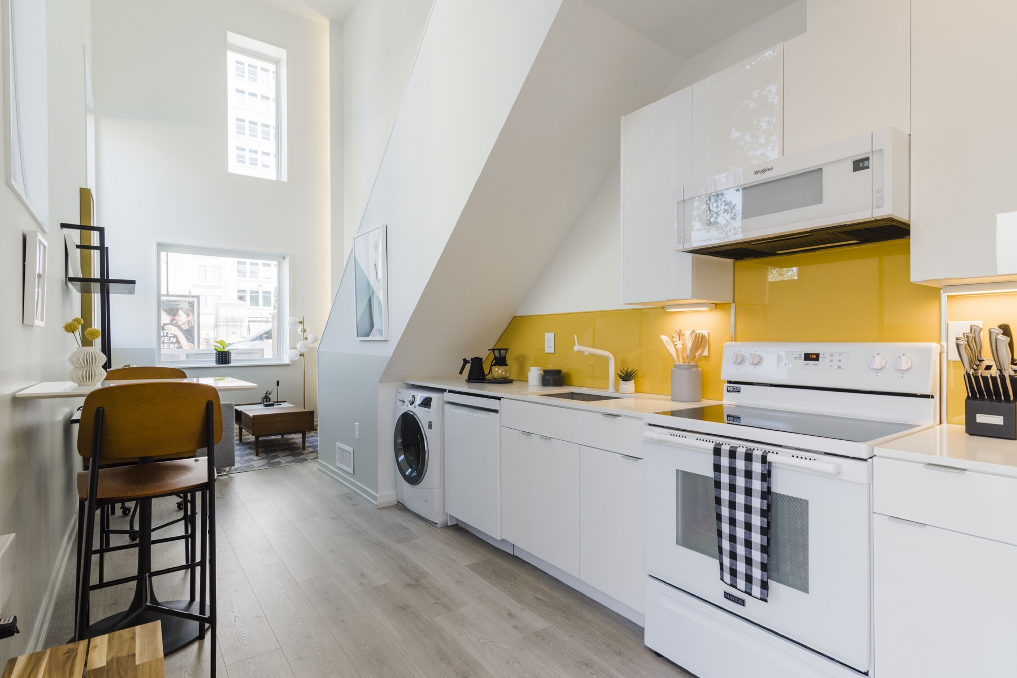 A modern kitchen with white appliances, a yellow accent wall, and a dining area with a bar stool.