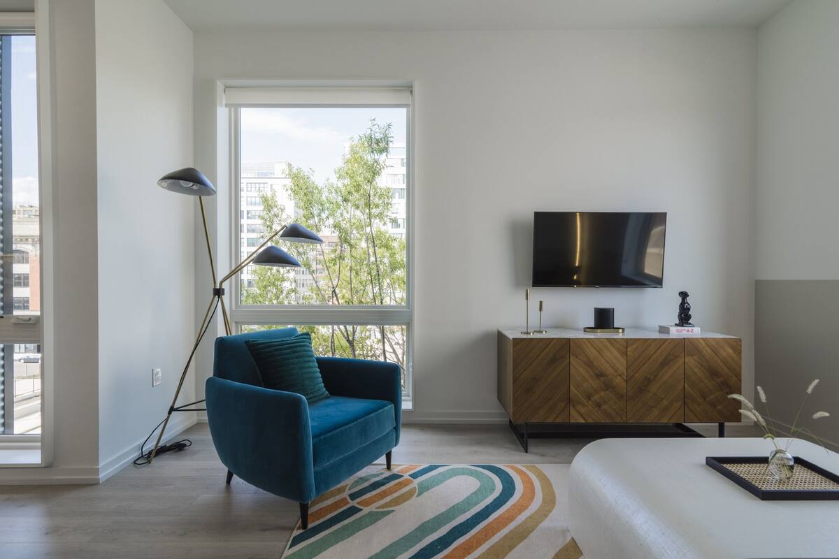 A modern bedroom with a large window, a blue armchair, a wooden cabinet, and a flat-screen TV.