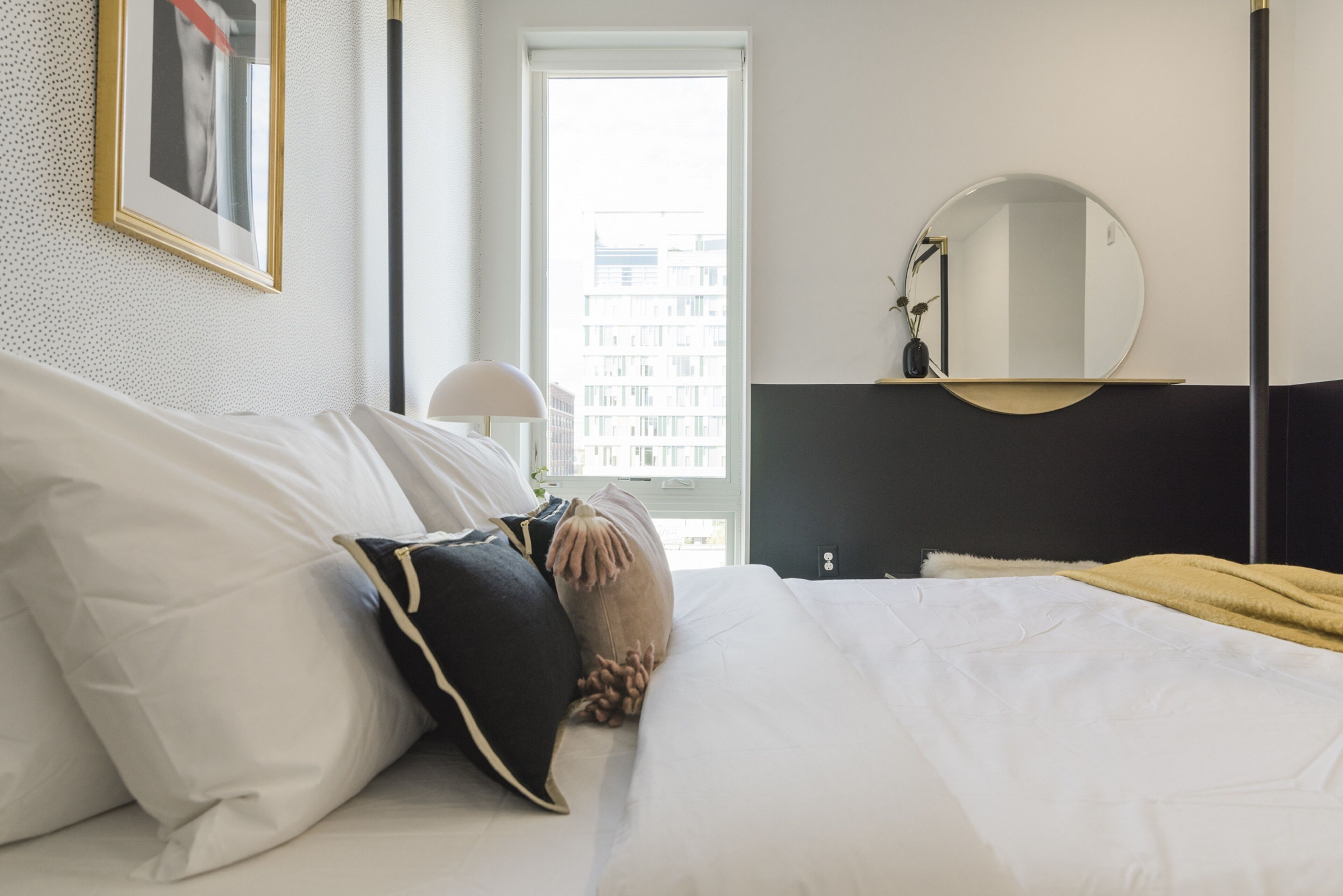 A hotel room with a neatly made bed, a round mirror, a window with a view of buildings, and a framed picture on the wall.