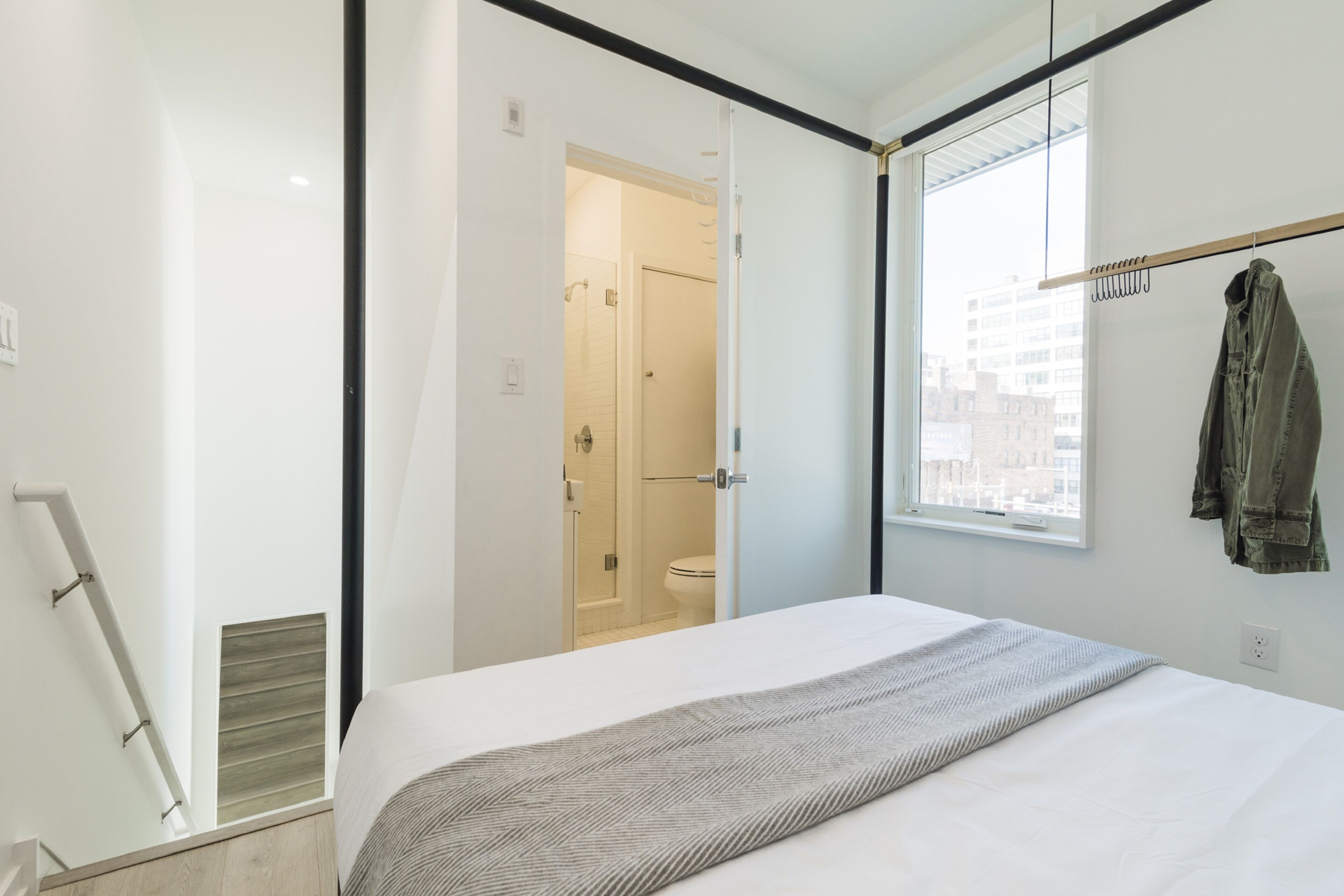 A modern bedroom with a large bed, a window with a view of buildings, and a wooden coat rack.