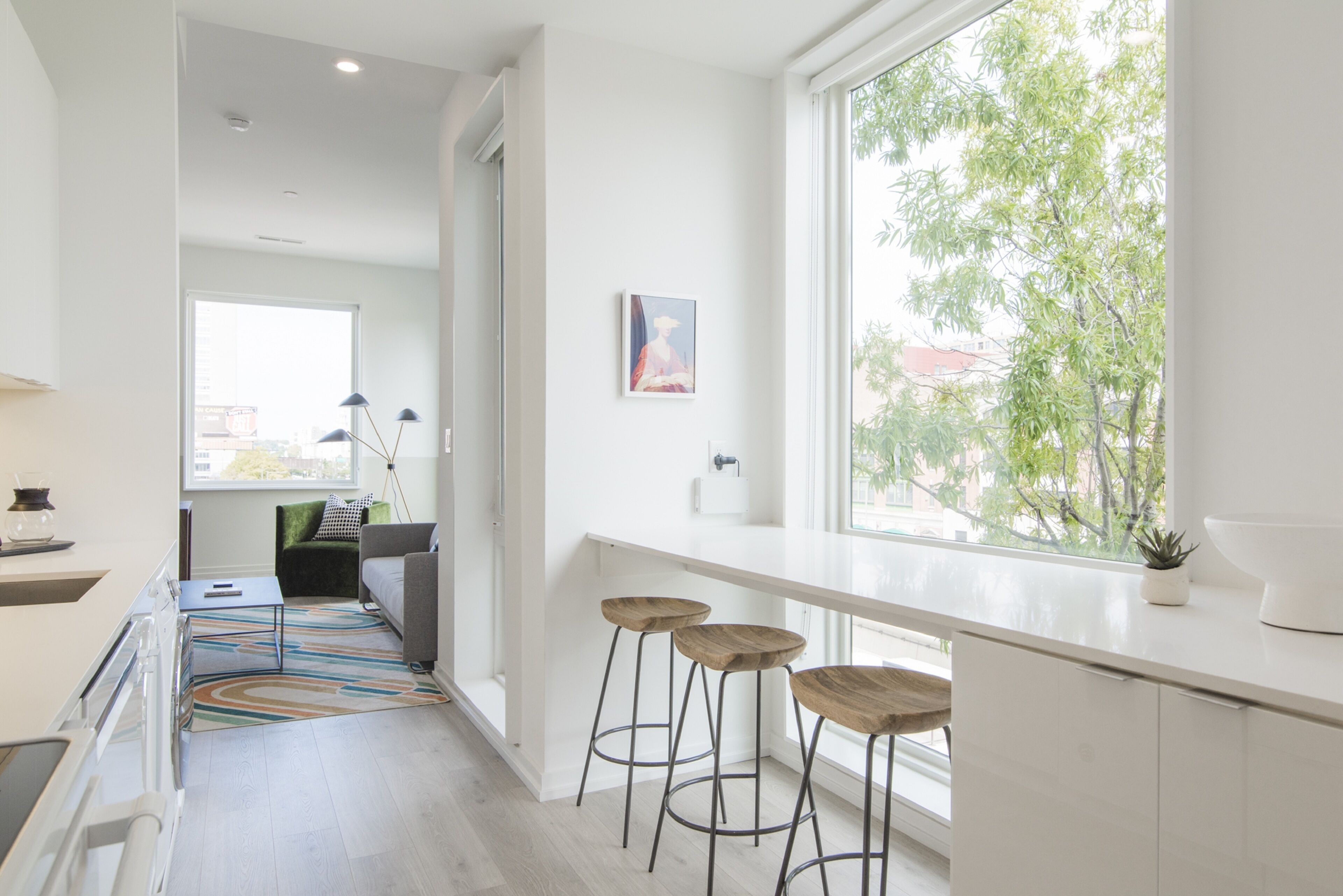 A modern kitchen with a long countertop, two stools, and a window with a view of trees.