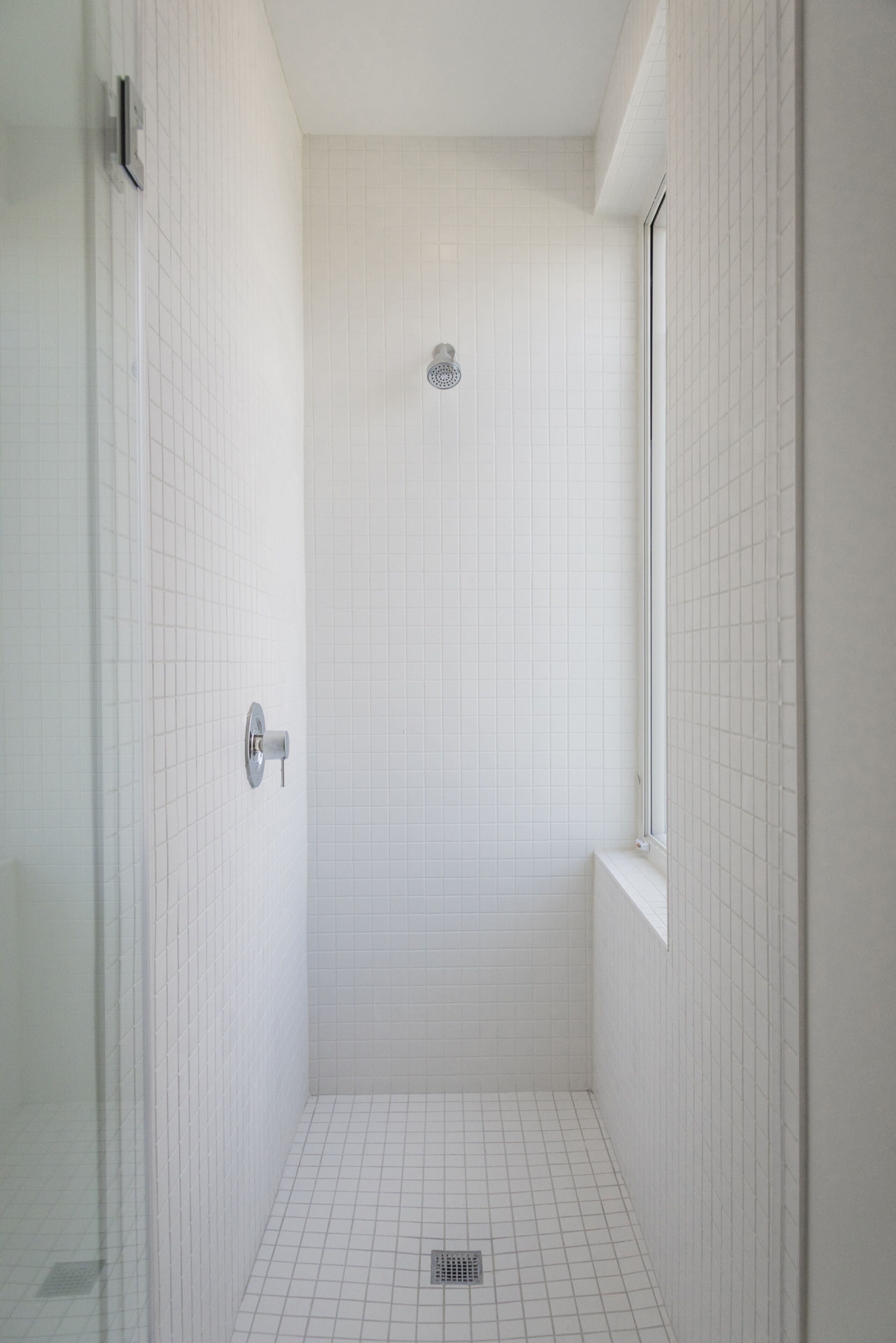 Une petite salle de douche aux murs recouverts de carreaux blancs, avec une fenêtre et une porte.