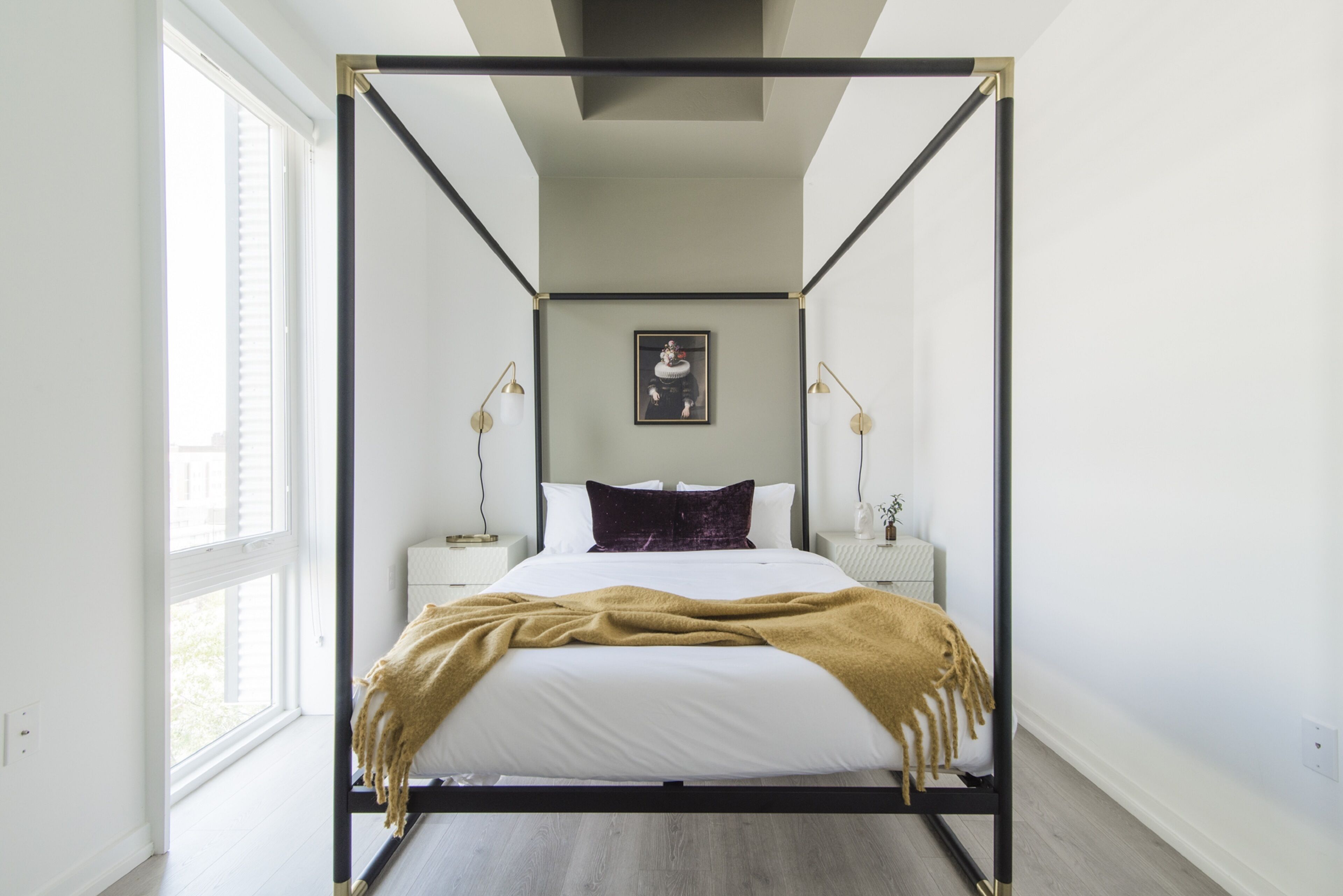 Un llit amb cortines i quatre pilars, amb una combinació de colors blancs i daurats, un quadre al damunt del llit i làmpades a cada costat.