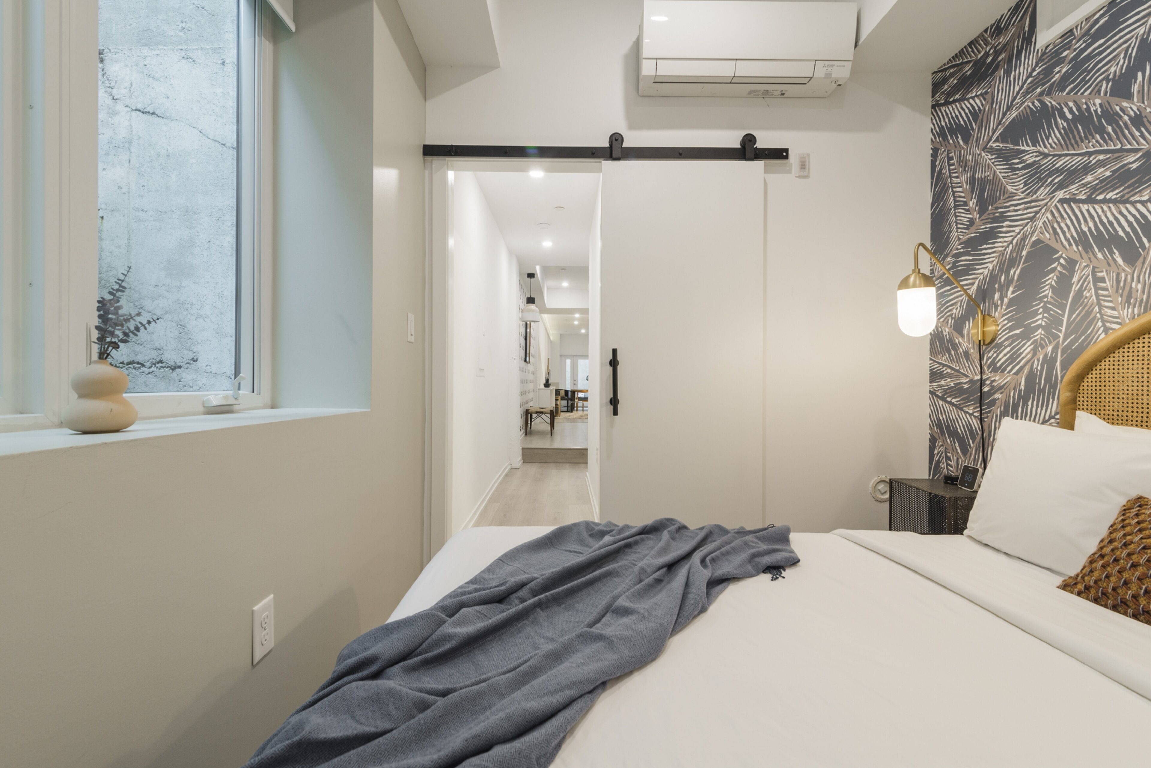 Une chambre moderne avec un lit, une fenêtre, un climatiseur fixé au mur et une porte donnant sur une autre pièce.