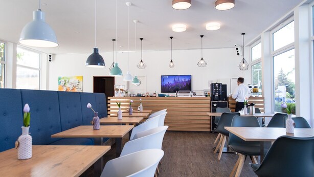 A modern cafe with wooden tables and blue chairs, a counter with a coffee machine, and a TV on the wall. - 🏨 SOVA Guesthouse Kassel 🌾 own hotel for up to 18 people (Kassel)