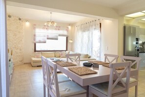 Un coin repas avec une table et des chaises blanches, un canapé et une cuisine en arrière-plan.