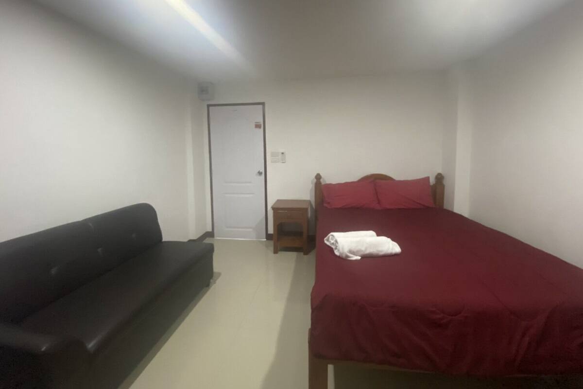 A bedroom with a bed, a red bedspread, a black sofa, and a white door.