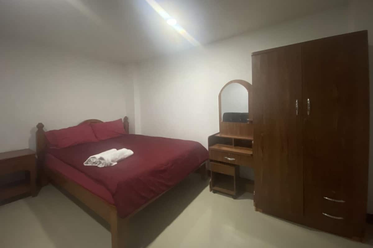 A bedroom with a bed, a wooden wardrobe, a mirror, and a small wooden dresser.