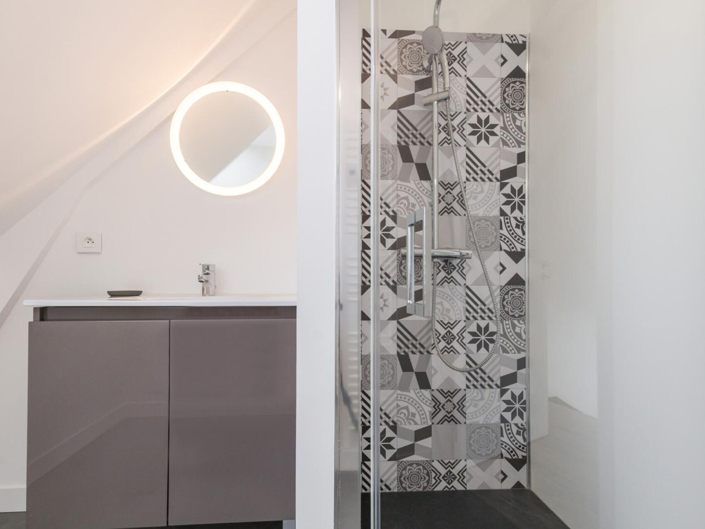 A modern bathroom with a geometric patterned shower wall, a round mirror with integrated lighting, and a minimalist vanity with a dark cabinet.
