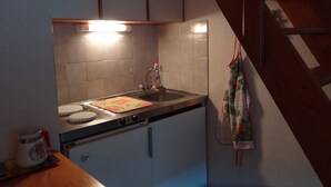 Una pequeña cocina con un lavamanos, una tetera y una estufa. Se observa una escalera de madera al fondo.