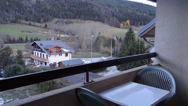 Un balcón con mesa y sillas, con vista a una casa y un valle boscoso.