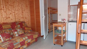 Una habitación pequeña y acogedora con suelo de madera, un sofá estampado, una mesa de madera con hervidor y un refrigerador pequeño.