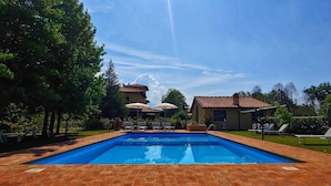 Une piscine rectangulaire bordée de carreaux, entourée de chaises longues et de parasols, avec des arbres et des bâtiments en arrière-plan.