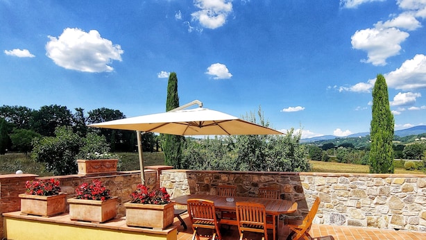 Une terrasse extérieure avec un mur en pierre, une table en bois, des chaises et un grand parasol. Des plantes en pot aux fleurs rouges et de grands arbres se trouvent en arrière-plan.
