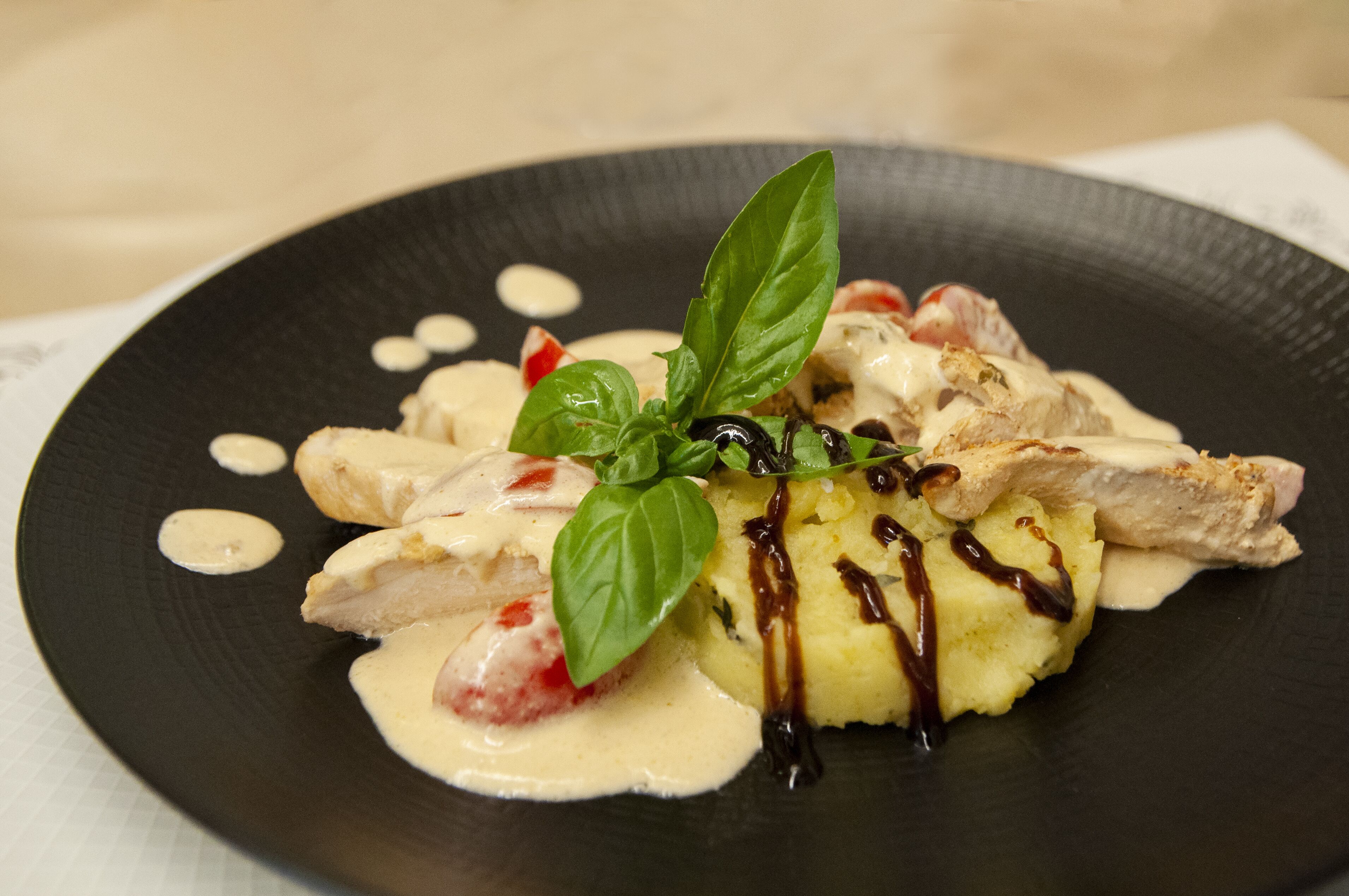 Une assiette de nourriture composée de poulet, de purée de pommes de terre et d’une sauce crémeuse, le tout garni de basilic et agrémenté d’un filet de sauce.