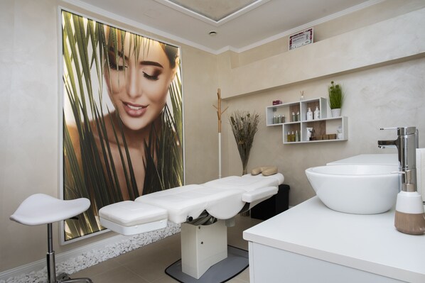 A modern spa room with a massage table, a large window with a view of palm leaves, a white sink, and a shelf with toiletries.