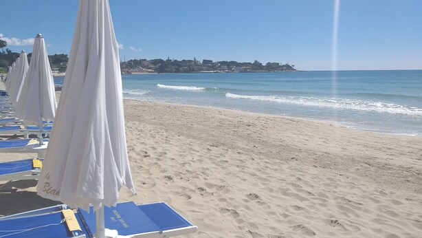 A beach with rows of white umbrellas and blue lounge chairs, a clear sky, and a distant view of a town by the sea. - Villa Flavia, Mediterranean charm. Apartment, private pool at100 mt sea (Fontane Bianche)