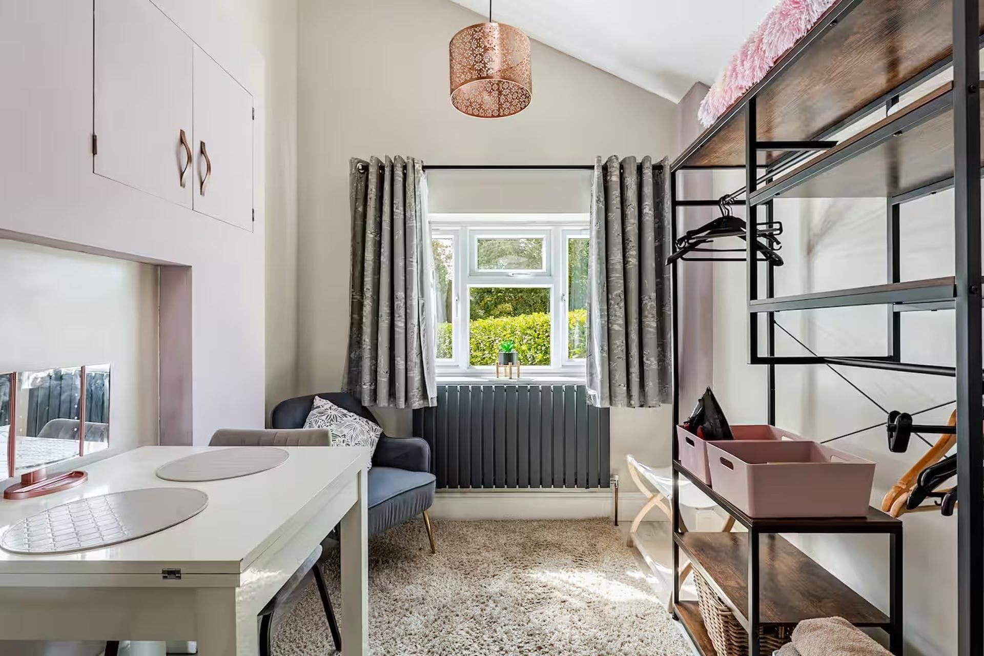Una habitación moderna con una mesa blanca, un sofá gris, una ventana con cortinas, un radiador y un perchero con cajas de almacenamiento.