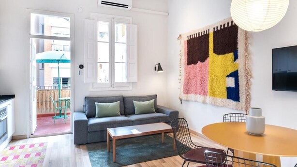 A modern living room with a grey sofa, a wooden coffee table, and a round dining table with wire chairs. There is a colorful rug, a large abstract wall hanging, and a TV mounted on the wall. - Oasis with a sunny terrace in the heart of Gracia (Barcelona)