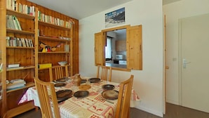 Ein Essbereich mit einem für vier Personen gedeckten Tisch, umgeben von Holzstühlen. Eine Bücherwand ist links zu sehen. In der Mitte steht ein Holzkasten mit einem Fenster, das in eine Küche blickt.