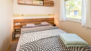 Un dormitorio con cabecera de madera, cama con ropa de cama de lunares, mesita de noche, estante sobre la cama y ventana con cortinas.
