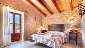Un dormitorio con pared de piedra, vigas de madera, una cama con colcha estampada, dos lámparas de noche y una mesita de noche de madera.