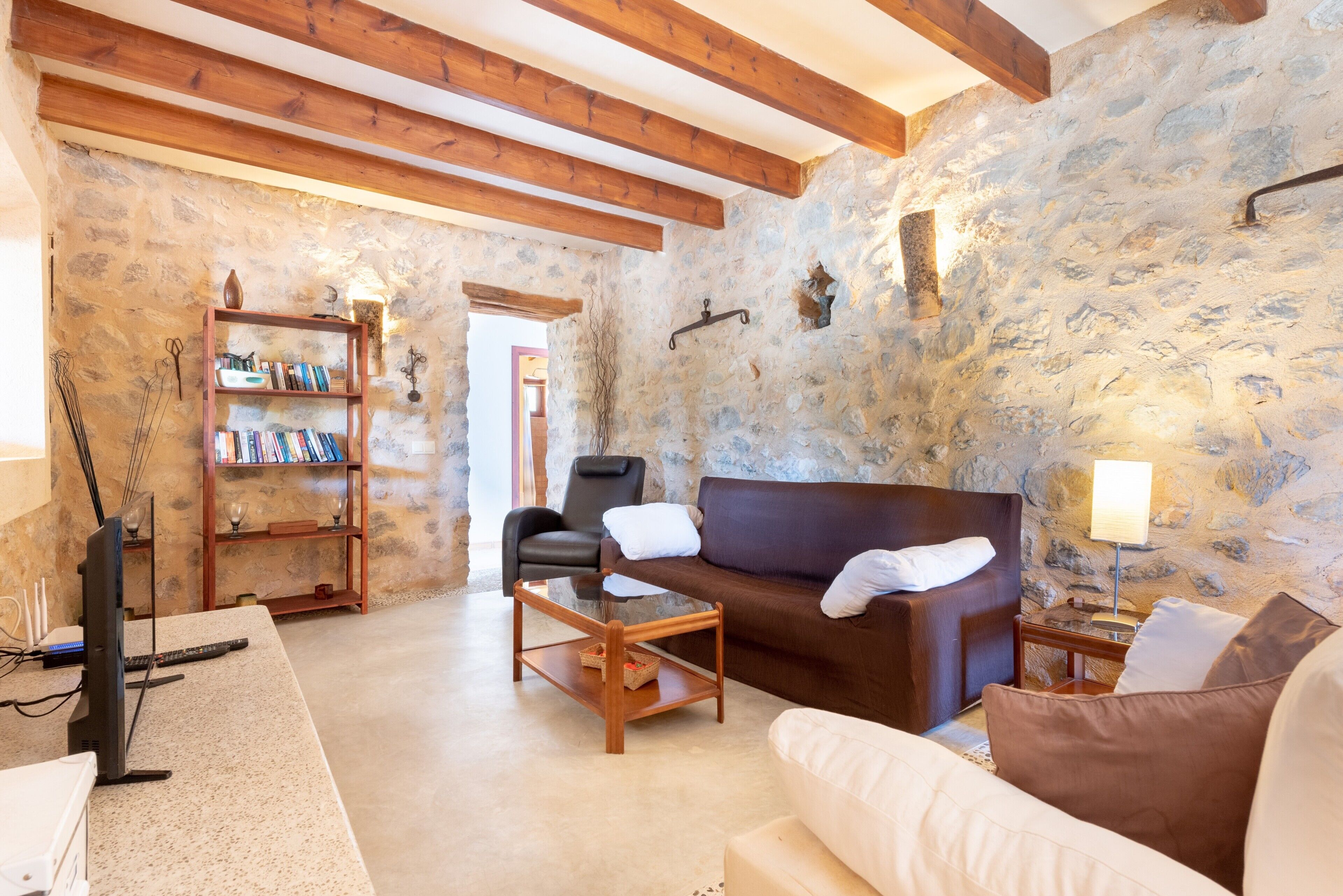 Une pièce avec un mur en pierre, des poutres apparentes, un canapé marron, un canapé blanc, une table basse en bois, une bibliothèque et un téléviseur.