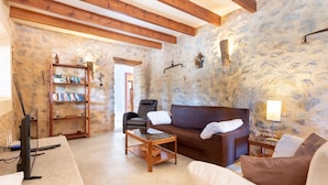 Una habitación con pared de piedra, vigas de madera, un sofá marrón, un sofá blanco, una mesa de centro de madera, una estantería y un televisor.