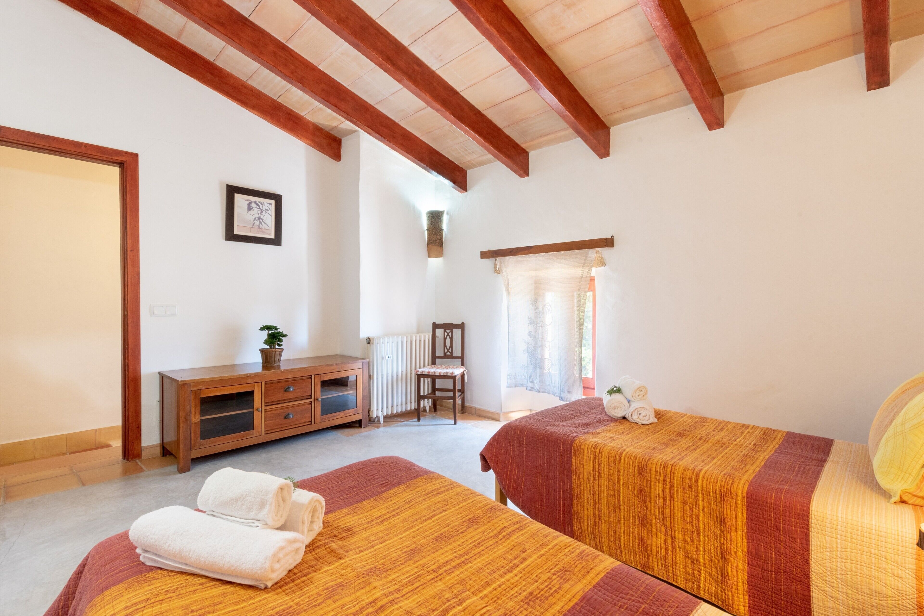 A hotel room with two beds, wooden beams, a radiator, a chair, a cabinet, and a window with curtains.