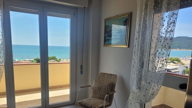 A room with a wicker chair, a framed picture, and a view of the sea through large glass doors.