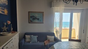 A room with a sofa, a dining table with a leaf-patterned tablecloth, a painting on the wall, and a view of the sea through a window.