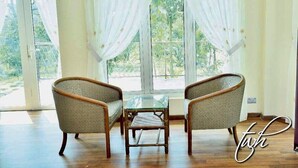 Una habitación con dos sillones de mimbre y una pequeña mesa de vidrio, ventanas amplias con cortinas traslúcidas y vistas a zonas verdes.