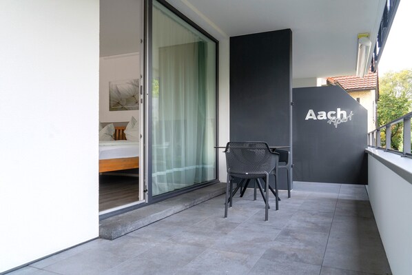 A modern balcony with a dining area, a sliding glass door leading to a bedroom, and a wall with the name 'Aach' visible.