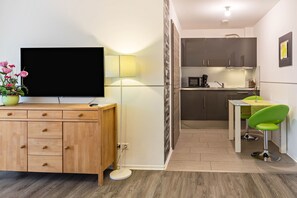 A modern room with a wooden cabinet, a flat-screen TV, a green chair, a small table, and a kitchen area.